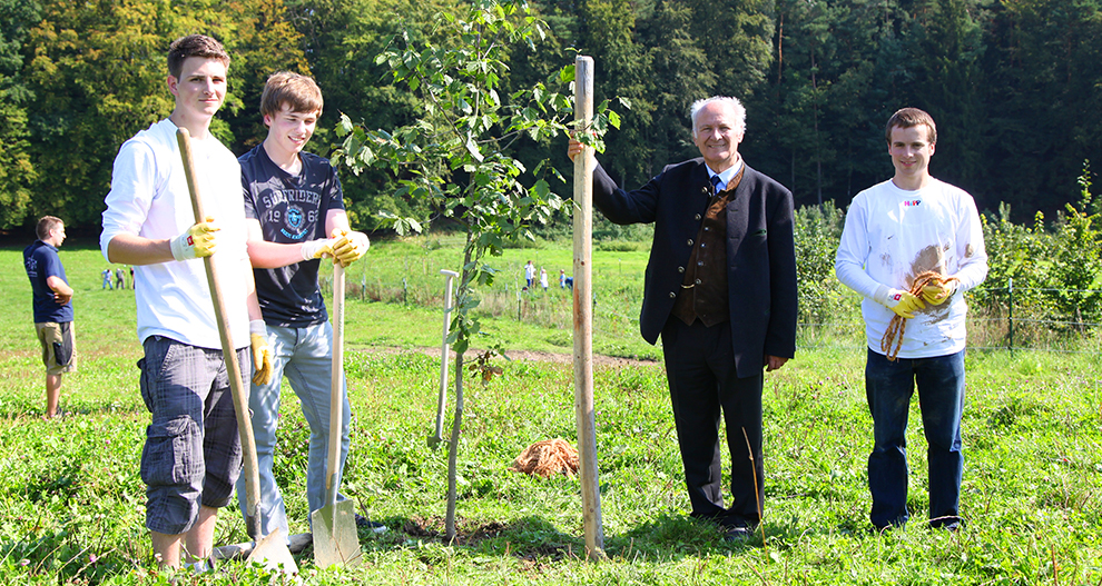 Four individuals are engaged in a tree planting activity in a grassy field with a backdrop of trees.