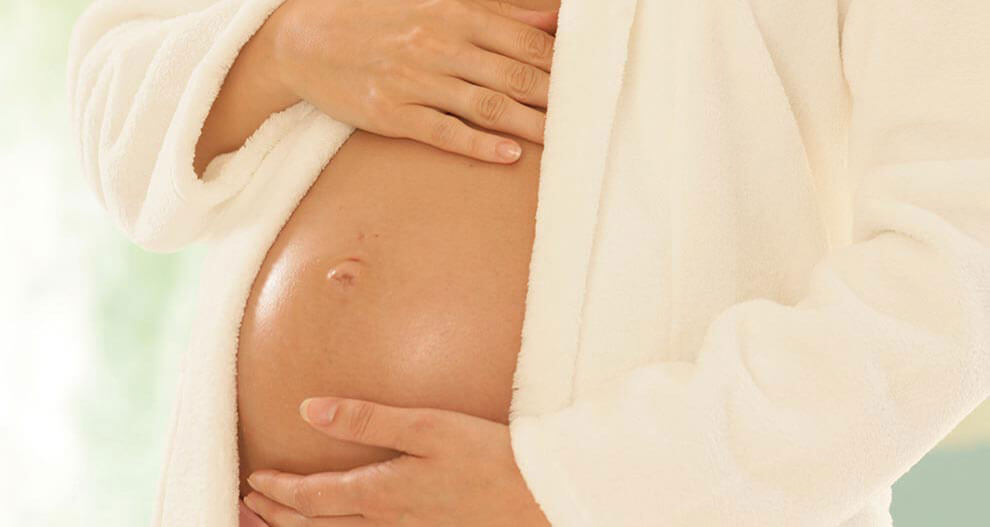 A pregnant woman in a white robe, holding her belly.