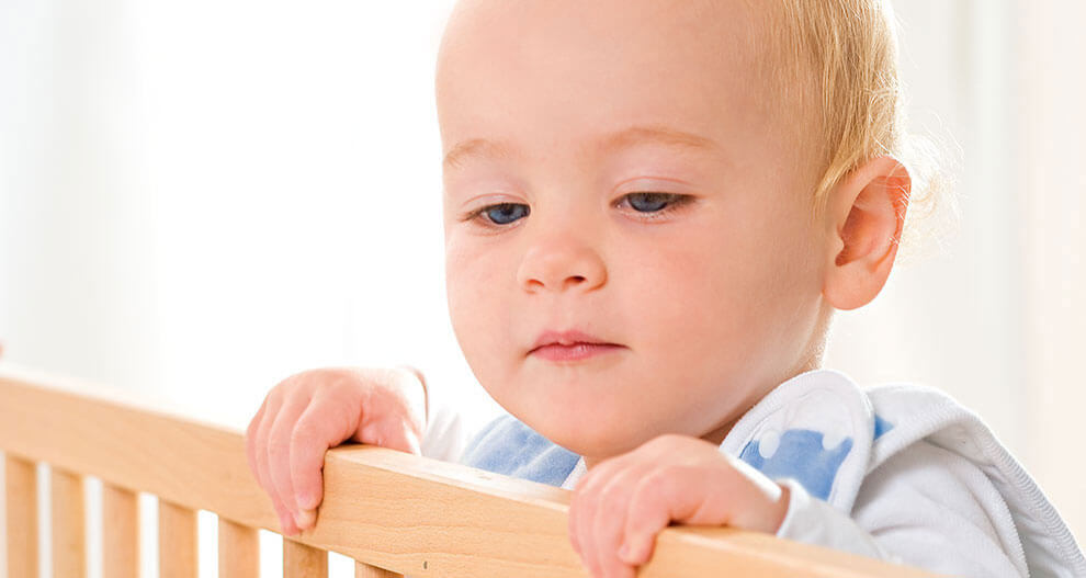 A baby with light hair is peering over a wooden crib railing.