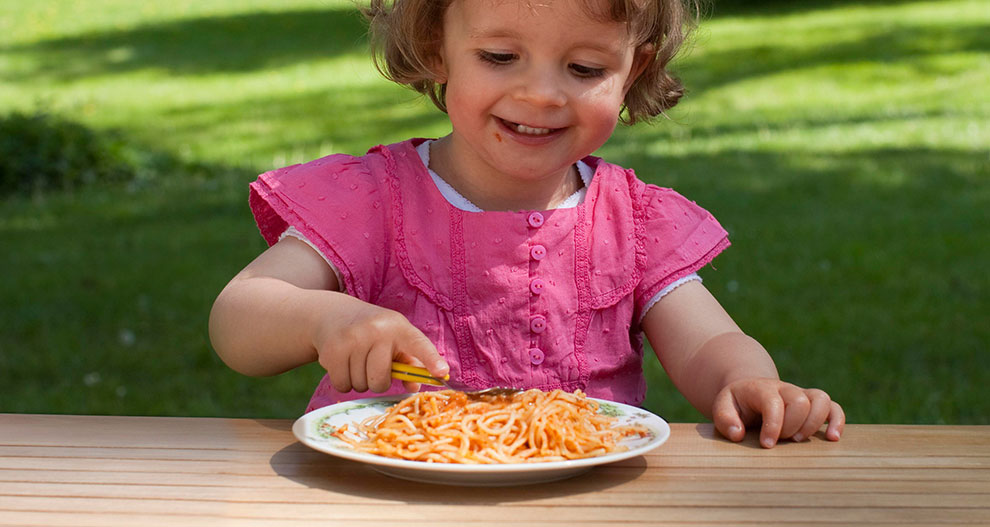 A child in a pink dress is eating spaghetti from a plate outdoors.