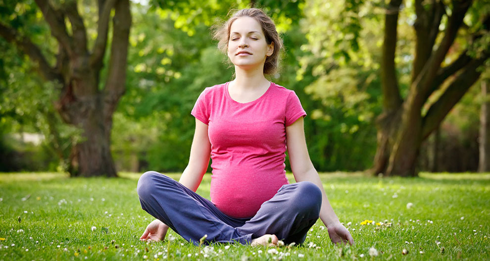 A woman in a pink shirt and blue pants sits cross-legged on grass, eyes closed, in a park with trees and flowers.