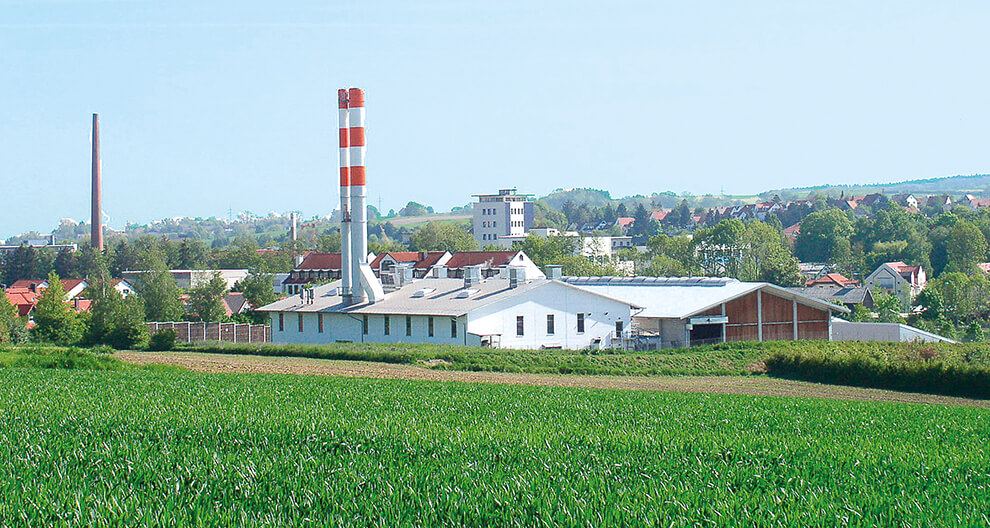A factory with a tall chimney stands in a green field, overlooking a town with red-roofed houses and a tall tower.
